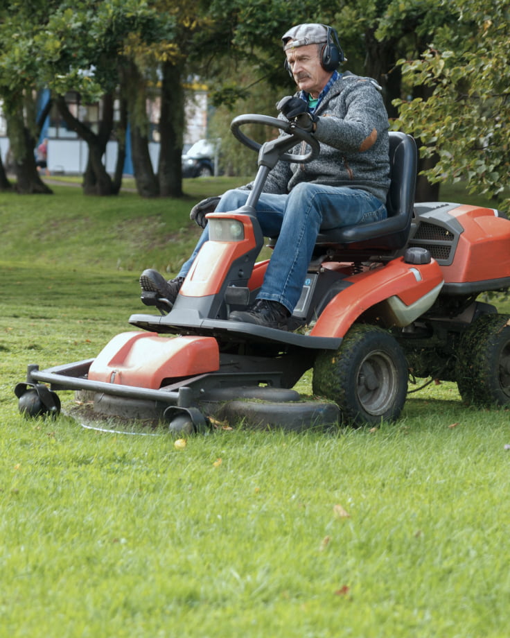 Older man wearing headphones drives orange lawn mower on grass.